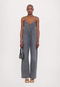 Combinaison en denim gris avec un décolleté en V plongeant et des bretelles fines, présentant des jambes larges. Associée à un petit sac à main noir en cuir crocodile et des chaussures à plateforme en bois.