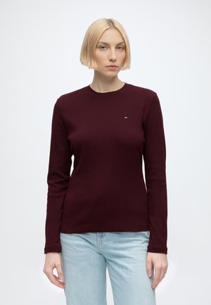 Femme aux cheveux blonds courts portant un chemisier à manches longues bordeaux foncé et un jean bleu clair sur un fond blanc uni.