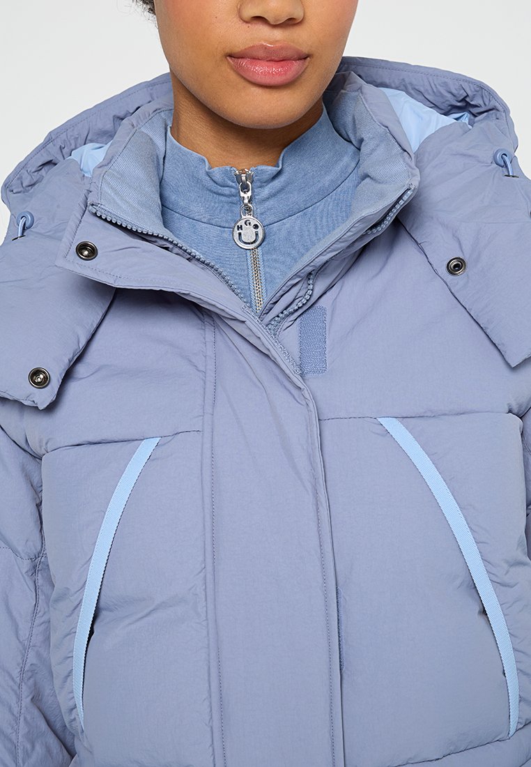 Donna che indossa un giubbotto imbottito di un leggero colore viola con colletto alto con zip e dettagli con bottoni a pressione, dotato di due tasche frontali con bordo azzurro.