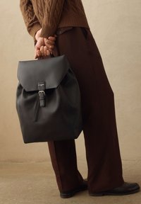 Mochila de cuero negro con solapa plegable y cierre de hebilla. Presenta una textura suave y una silueta redondeada, complementada con pantalones marrones.