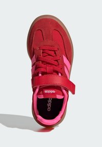 Röd och rosa sportskor med övre del i mocka, tygskosnören, en kardborreband och en brun gummisula. Logotypen är placerad på tungan.