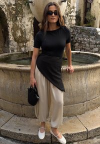 Top noir ajusté avec un ourlet asymétrique, pantalon large crème, ballerines blanches et un petit sac à main noir près d'une fontaine en pierre.