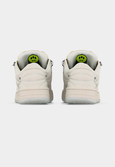 Ζευγάρι εκρού αθλητικών παπουτσιών από πίσω, με πράσινες ετικέτες στη γλώσσα που φέρουν μαύρα σχέδια τριών τελειών, σε απλό φόντο.