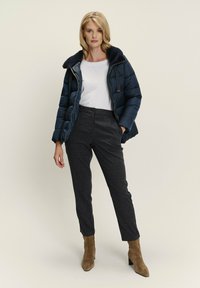 Frau steht da in einer marineblauen Daunenjacke mit Pelzkragen, weißem Hemd, dunklen karierten Hosen und braunen Wildleder-Stiefeletten.