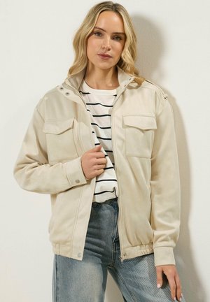 Kobieta o blond włosach, ubrana w beżową kurtkę zapinaną na suwak, na białej i czarno paskowanej koszulce oraz w jasnoniebieskie dżinsy, stojąca na tle jednolitej ściany.