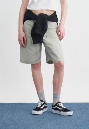 Isik, kes kannab helehallid põlvpikkused lühikesed püksid, mustad tossud valgete detailidega, hallid sokid, valge lühike topp ja vööst seotud musta dressipluusi.