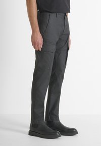 Uomo che indossa pantaloni cargo neri con tasche laterali e stivali neri alla caviglia, in piedi contro uno sfondo bianco semplice.