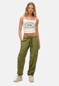 Groene cargo pants met meerdere zakken, elastische boorden en een trekkoord in de taille. Draag een witte tanktop met groene tekst.