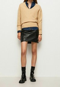 Pull en mélange de laine beige en v, superposé sur une chemise en denim foncé, associé à une jupe portefeuille en cuir noir et des bottines vernies.