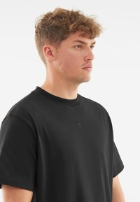 Černé tričko s krátkým rukávem, vyrobené z bavlny, má kulatý výstřih a subtilní reliéfní logo na hrudi. Hladká textura.