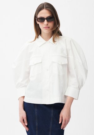 Frau trägt ein übergroßes weißes Hemd mit Puffärmeln, dunkle Sonnenbrille und einen dunkelblauen Jeansrock vor einem einfarbigen Hintergrund.