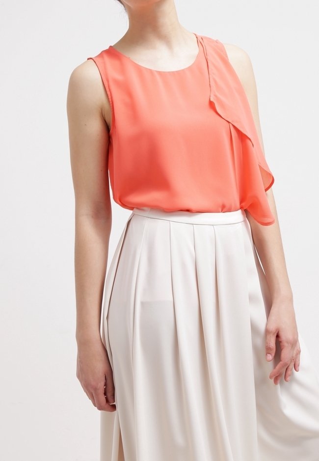 Femme portant un chemisier corail sans manches et une jupe plissée ivoire taille haute, debout devant un fond blanc.