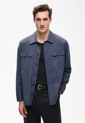 Junger Mann trägt eine blaue Hemdjacke über einem schwarzen Hemd und schwarzen Hosen mit Gürtel, stehend vor weißem Hintergrund.