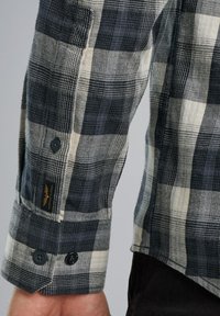 Flanellen overhemd met zwart, grijs en crème ruitpatroon; beschikt over manchet met twee zwarte knopen; geweven stoftextuur; merketiket op de mouw.