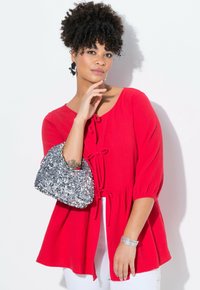 Top rosso testurizzato con maniche a tre quarti e lacci frontali, abbinato a una borsa d'argento con paillettes, su uno sfondo bianco.