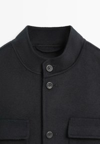 Cappotto di lana nero con colletto alto, due tasche frontali e quattro bottoni sul davanti. Il tessuto ha una texture liscia e morbida.