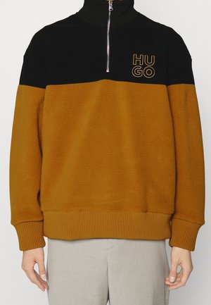Puoli-vetoketjullinen fleece-pullover mustalla yläosalla, sinappivärisellä vartalolla, resorilla hihoissa ja helmassa, sekä "HUGO"-logo vasemmassa rinnassa sinappivärisellä kirjonnalla.