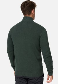 Sötétzöld kötött pulóver magas gallérral, bordázott mandzsettákkal és aljjal. A textúra sima, enyhén nehéz megjelenésű.