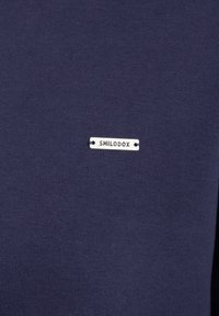 T-shirt blu navy con una texture liscia e un piccolo etichetta argentata che riporta il testo "SMILODOX," attaccata con due occhielli metallici.