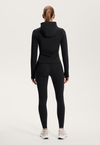Černá přiléhavá sportovní bunda s kapucí, spárovaná s odpovídajícími černými legínami. Design má elegantní linie a hladkou strukturu.
