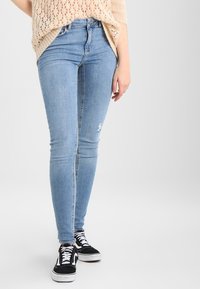 Smala ljusblå jeans med hög midja, med slitna detaljer vid knäna, tillsammans med svarta och vita sneakers och en stickad beige topp.