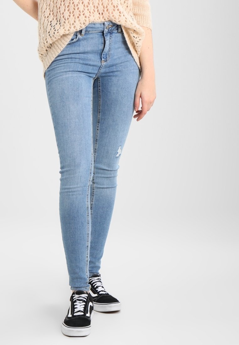 Smala ljusblå jeans med hög midja, med slitna detaljer vid knäna, tillsammans med svarta och vita sneakers och en stickad beige topp.