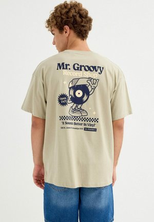 Joven con cabello rizado que lleva una camiseta beige con el personaje y texto de "Mr. Groovy Record Shop" promocionando discos de vinilo.