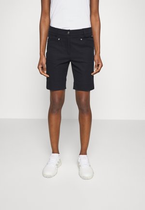 Persona que lleva pantalones cortos negros hasta la rodilla, camiseta blanca sin mangas y zapatillas deportivas blancas, de pie sobre un fondo claro y liso.