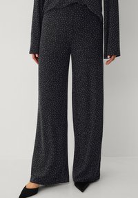 Schlaghosen aus schwarzem Stoff mit weiß gesprenkeltem Muster; weiche Textur, elastischer Bund; kombiniert mit schwarzen spitzen Schuhen.