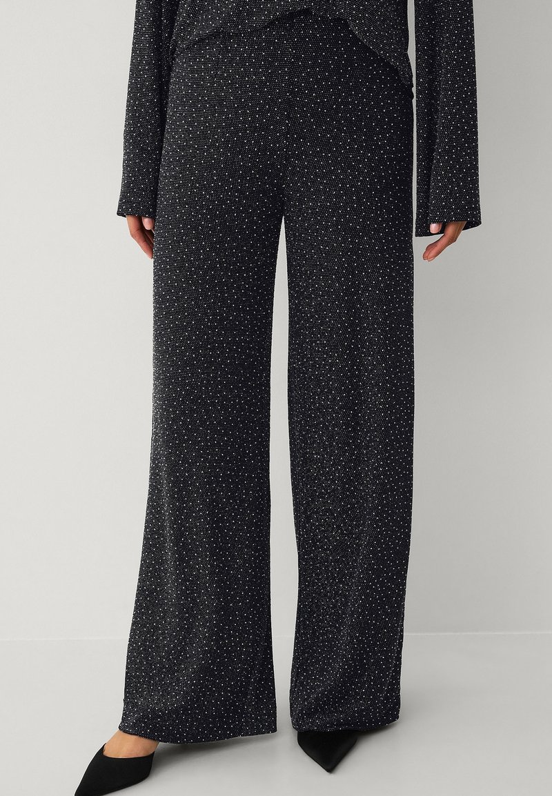 Schlaghosen aus schwarzem Stoff mit weiß gesprenkeltem Muster; weiche Textur, elastischer Bund; kombiniert mit schwarzen spitzen Schuhen.