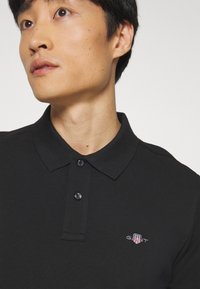 Tânăr cu păr negru scurt purtând un tricou polo negru cu un mic logo multicolor GANT pe piept, uitându-se spre stânga.
