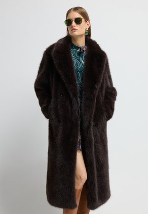 Brun faux fur frakke med dobbeltradet design, stort krave og sidelommer. Modellen har et mønstret kjole og tilbehør på.