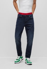 Vaqueros de mezclilla azul oscuro con corte recto, con una cinturilla de marca roja 'HUGO', combinados con zapatillas verdes y blancas.
