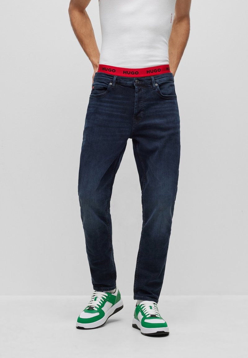 Vaqueros de mezclilla azul oscuro con corte recto, con una cinturilla de marca roja 'HUGO', combinados con zapatillas verdes y blancas.
