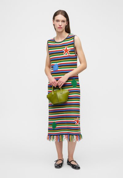 Frau in einem ärmellosen, gestreiften Midikleid mit meeresthematischen Aufnähern, die eine kleine grüne Handtasche hält und schwarze flache Schuhe mit Riemen trägt.