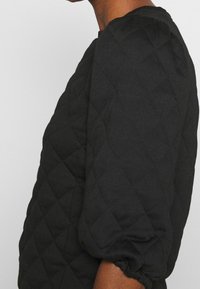 Svart quiltad topp med en texturerad mönstring, rund halsringning och puffärmar med elastiska manschetter, som visar en strukturerad silhuett.