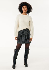 Krämfärgad stickad tröja, svartvitt rutigt blyertsskjol, svarta knähöga stövlar och genomskinliga svarta strumpbyxor, står framför en enfärgad bakgrund.