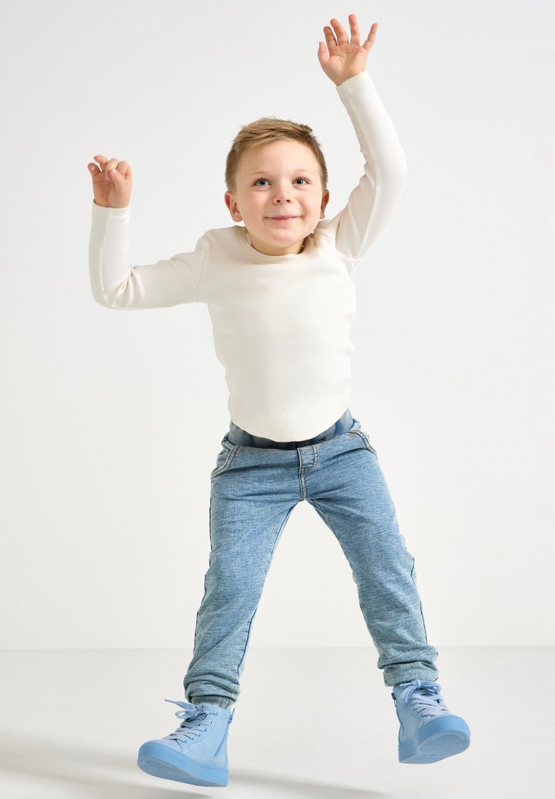 Hvit langermet skjorte, blå denim joggebukser og lyseblå høye sneakers. Barnet hopper med armene hevet mot en ensfarget bakgrunn.