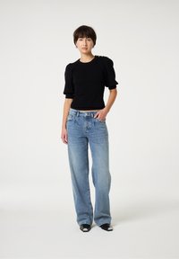 Zwarte ribgebreide top met pofmouwen, gecombineerd met lichtblauwe wijde jeans. Model draagt zwart-witte puntige schoenen. Minimalistische achtergrond.