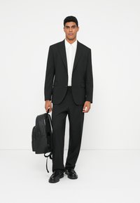 Traje negro con corte ajustado, camisa blanca y mochila negra. Tejido suave, solapas con muesca y pantalones rectos. Zapatos de cuero.