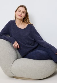 Navyblauer Strickpullover mit weitem Ausschnitt, langen Ärmeln und lässiger Passform. Weiche Textur. Trägt dazu passende navyfarbene Hosen auf einem strukturierten cremefarbenen Stuhl.
