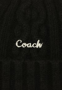 Black knit beanie featuring a ribbed texture and embroidered white "Coach" logo on the front. Soft, warm material suitable for cold weather.