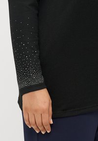 Μαύρο μακρυμάνικο τοπ με υφή. Οι μανσέτες διαθέτουν μια κλίμακα από ασημί �κρύσταλλα, προσθέτοντας μια διακριτική λάμψη.