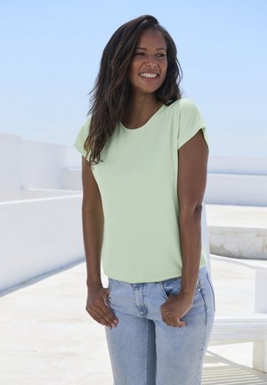 Hellgrünes, strukturiertes Oberteil mit kurzen Ärmeln und rundem Ausschnitt, kombiniert mit hellblauen Jeans. Steht vor einem hellen, outdoor Hintergrund.