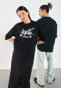 Svart oversized t-skjorte med hvit grafikk av Mickey og Minnie Mus med "Disney" tekst under. Modell bruker den med svart skjørt og lyse jeans.