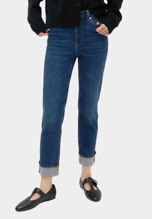 Person iført opkrogede mørkeblå jeans, sort knappet cardigan og sorte Mary Jane-sko med spænde, stående på en lys baggrund.