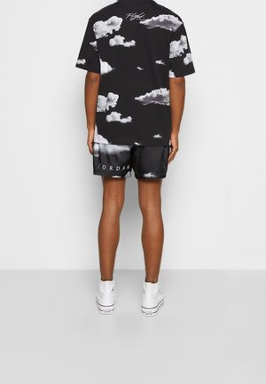 Persona che indossa una maglietta e pantaloncini con motivo a nuvole in bianco e nero con i testi "Flight" e "Jordan", in piedi di fronte a uno sfondo bianco uniforme.