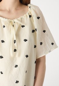 Ανοιχτό κρεμ μπλούζα με κοντά, φαρδιά μανίκια; διαθέτει μαύρη λουλουδάτη κεντητική και συγκεντρωμένο λαιμό με λεπτομέρεια σε σχήμα φιόγκου.