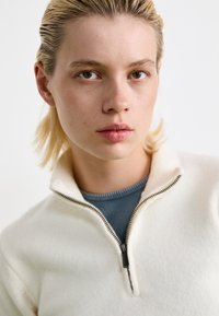 White zip-up fleece sweater with a high collar, featuring a black zipper and a textured surface, layered over a blue ribbed top.