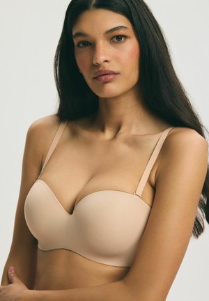 Woman with long dark hair wears a nude strapless bra, looking forward with a neutral expression on a plain light background.